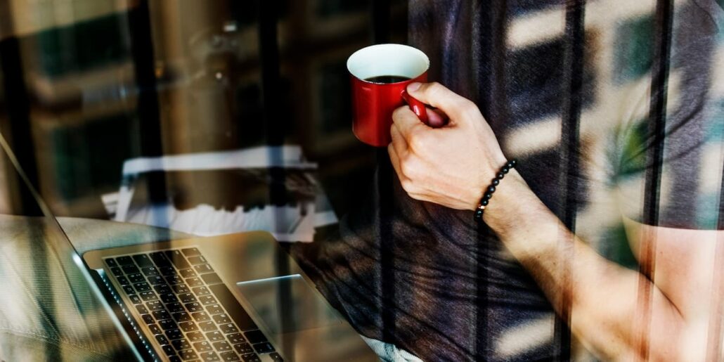 Karriere 8 Person mit roter Kaffeetasse in der Hand arbeitet an einem Laptop, Aufnahme durch ein Fenster mit Spiegelungen.