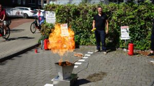 Ein Mann demonstriert im Freien den Umgang mit Feuer und Feuerlöschern, während eine große Flamme aus einem Metallbehälter lodert. Zuschauer beobachten die Vorführung.