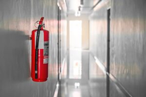 Roter Feuerlöscher an einer grauen Wand in einem hellen, langen Flur montiert.