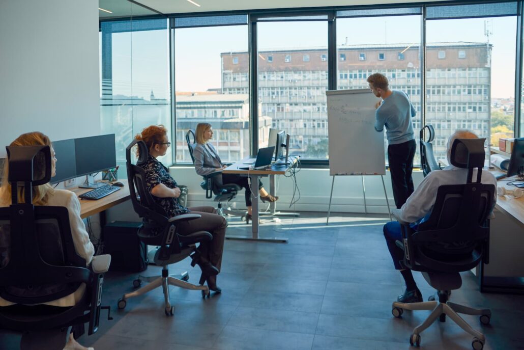 Karriere 5 Mitarbeiter sitzen in einem modernen Büro und folgen einer Präsentation an einem Flipchart vor großen Panoramafenstern.
